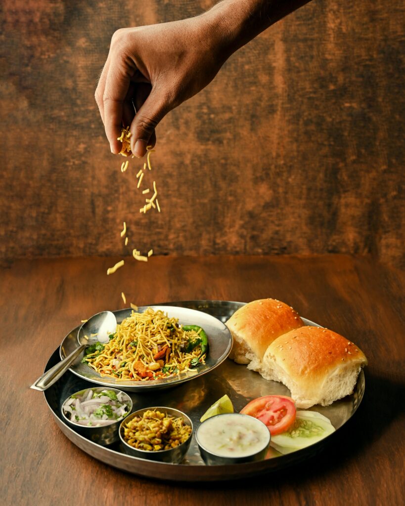 Home A close-up of misal pav, a spicy Indian dish, with toppings being added by hand.