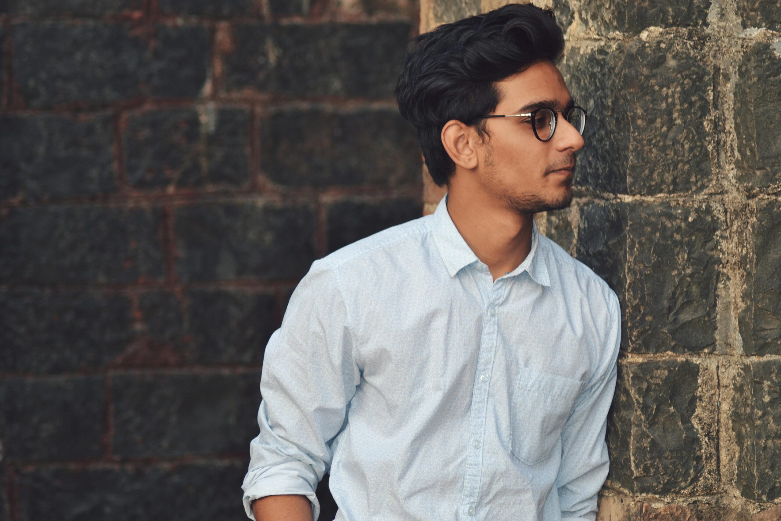 Testimonial A stylish young man in glasses leans against a brick wall wearing a casual light blue shirt.